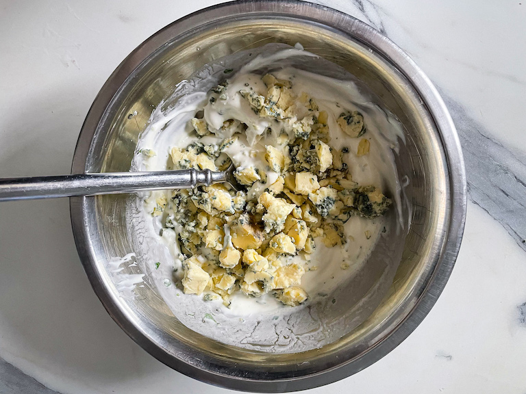 Adding blue cheese crumbles to a bowl of mayonnaise and sour cream to make blue cheese dip