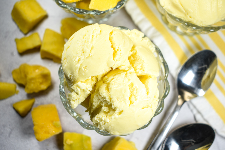 Horizontal shot of a bowl of mango ice cream, surrounded by spoons and chunks of mango