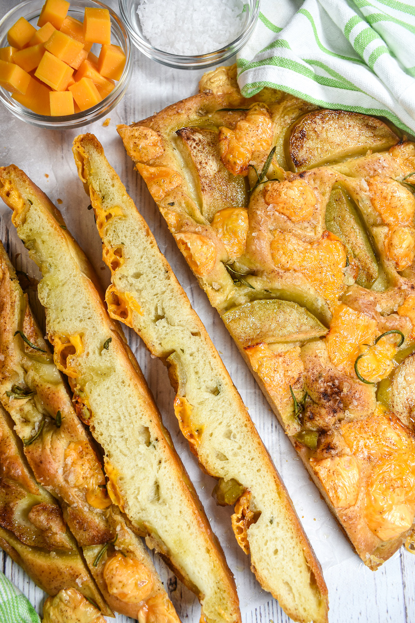 Apple focaccia sliced and arranged on a white surface with a bowl of salt and bowl of cheese cubes