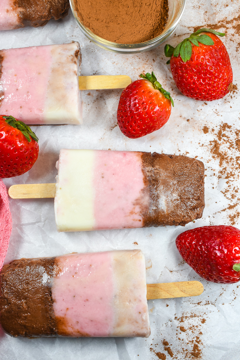 Neapolitan popsicles, strawberries, cocoa powder arranged on a white surface