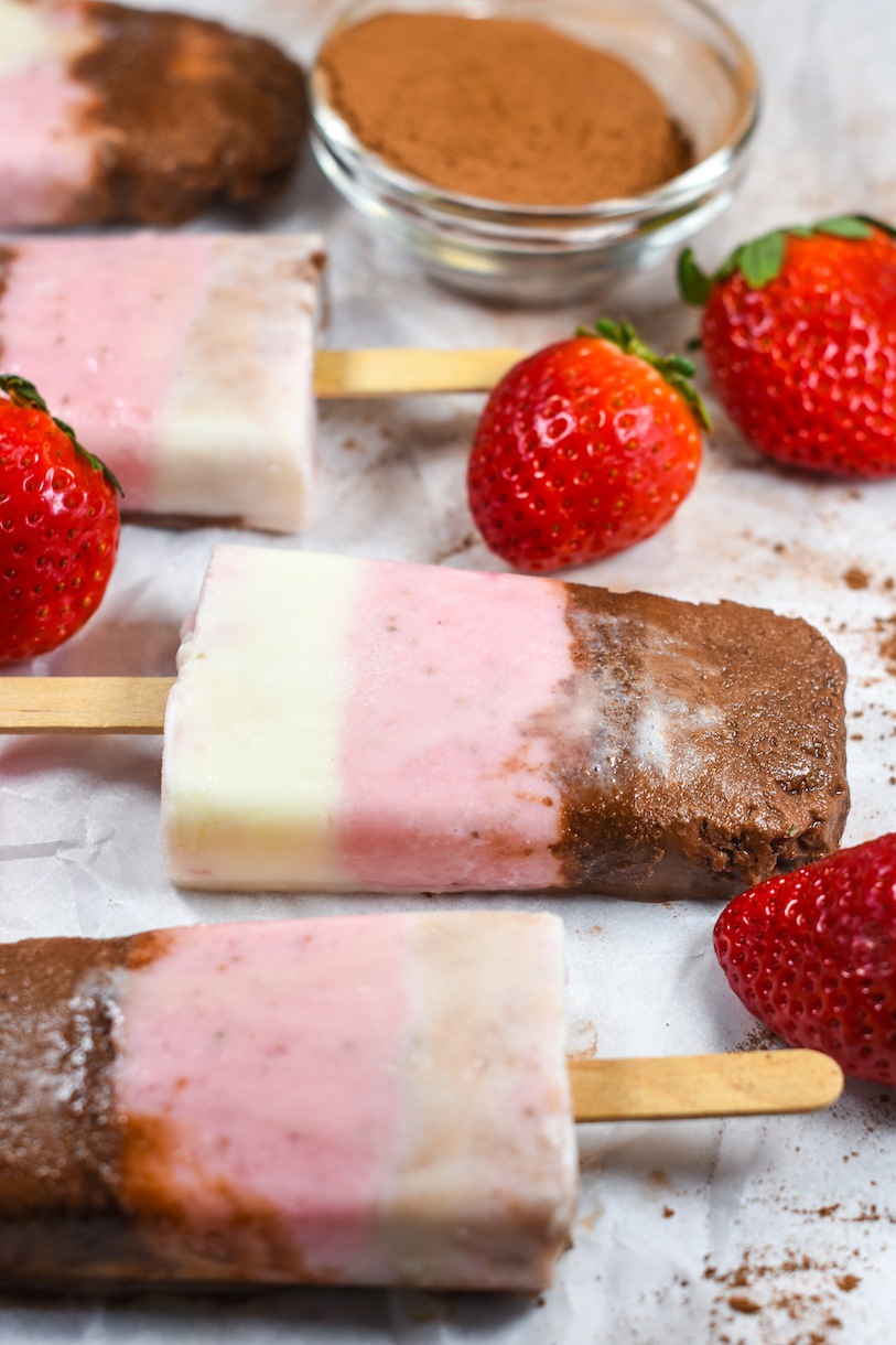 Neapolitan ice cream pops on a white surface with strawberries and cocoa powder