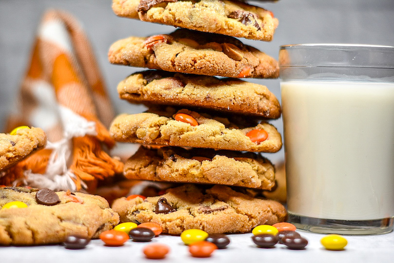 A stack of Reese's peanut butter cup cookies and a glass of milk surrounded by Reese's Pieces candies