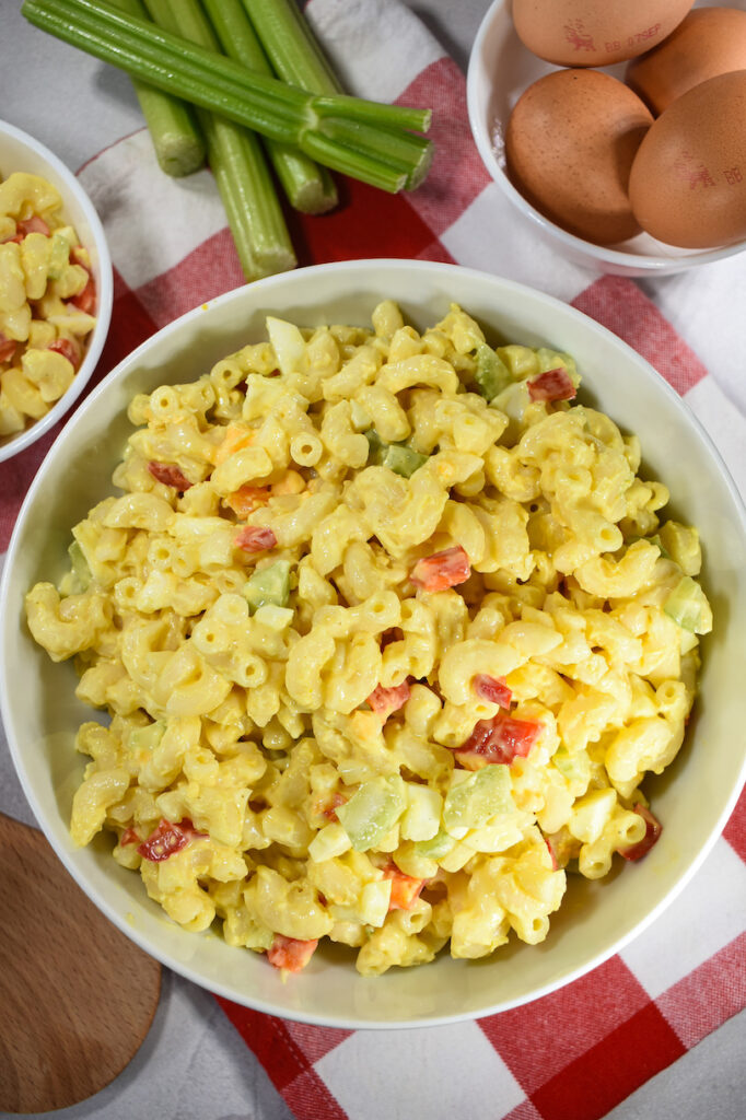 bowl of macaroni salad with a bowl of eggs and stalks of celery on a checkered towel