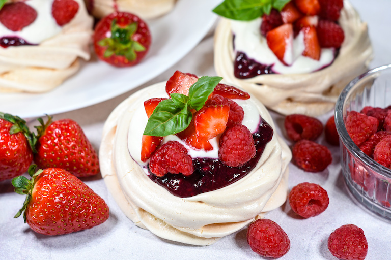 Horizontal shot of a miniature pavlova with compote, cream, and berries, surrounded by fresh raspberries and strawberries