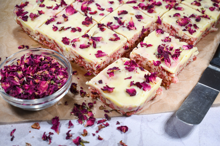 Dish of dried rose petals and squares of raspberry rose rice krispie treats on a sheet of parchment