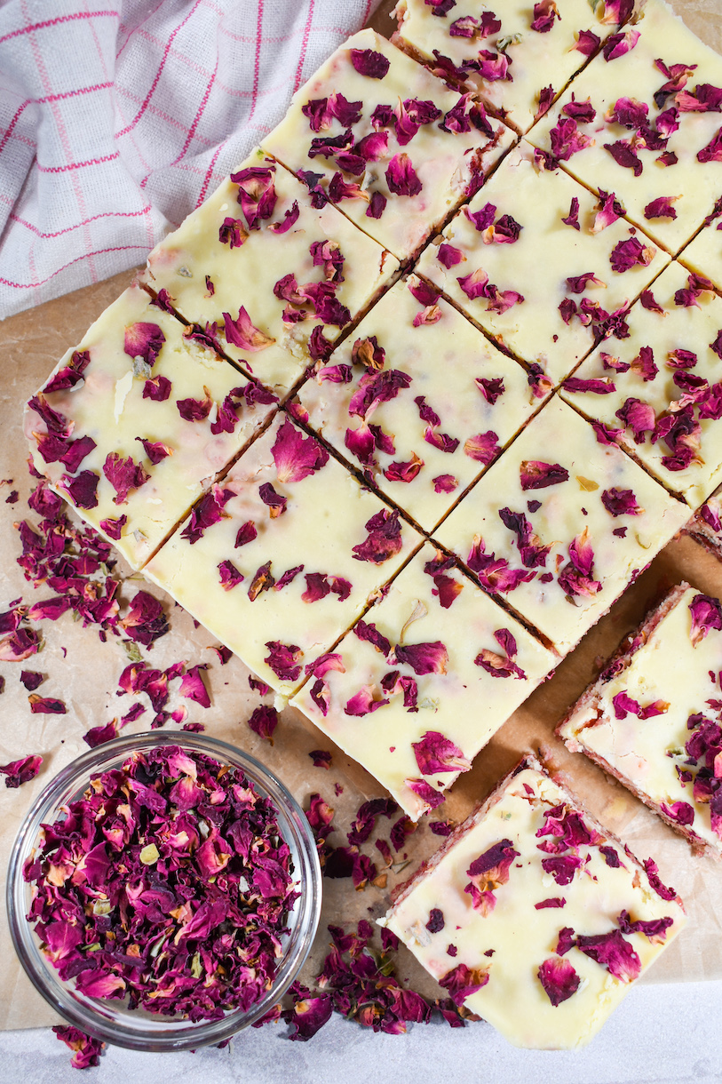 A slab of raspberry rose cereal treats cut into squares, surrounded by a plaid towel and a bowl of dried rose petals
