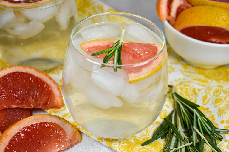 A glass of grapefruit water surrounded by grapefruit wedges and rosemary sprigs