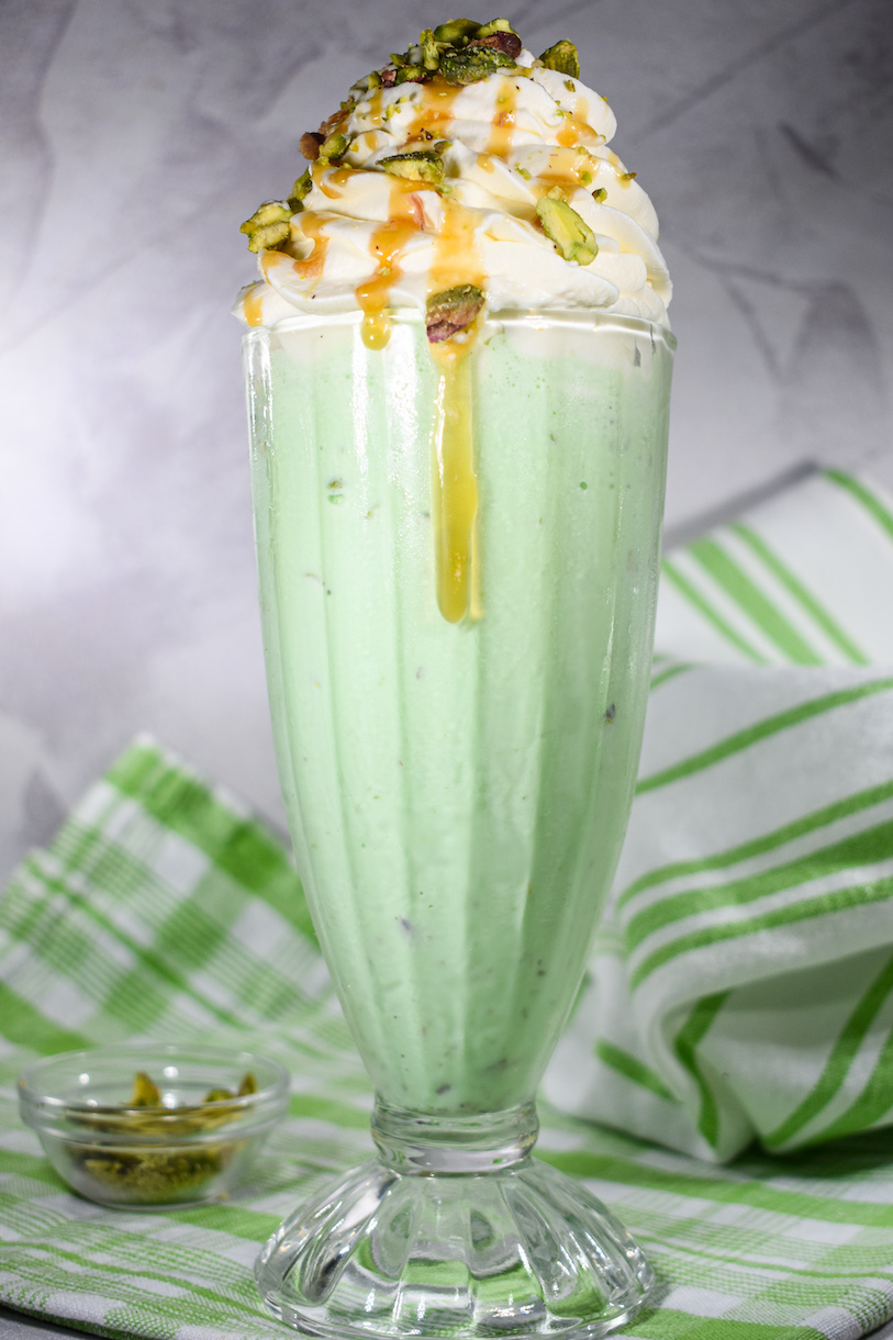 A pistachio milkshake in a tall glass with caramel dripping down the side