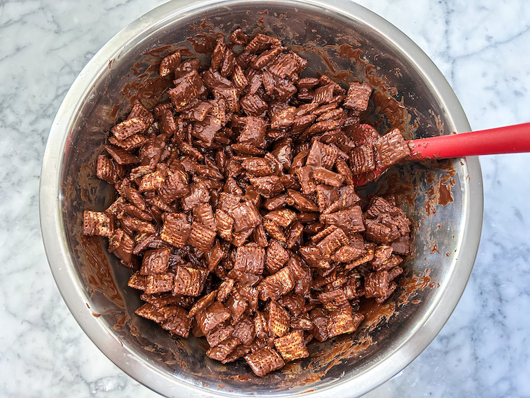 Cereal coated in chocolate, in a large metal bowl with a rubber spatula