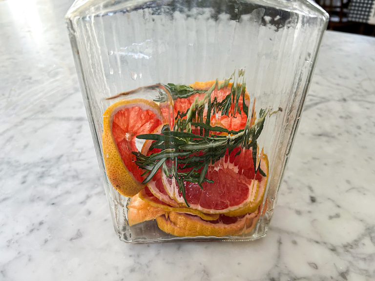 A glass jar containing slices of grapefruit and sprigs of rosemary, sitting on a marble countertop