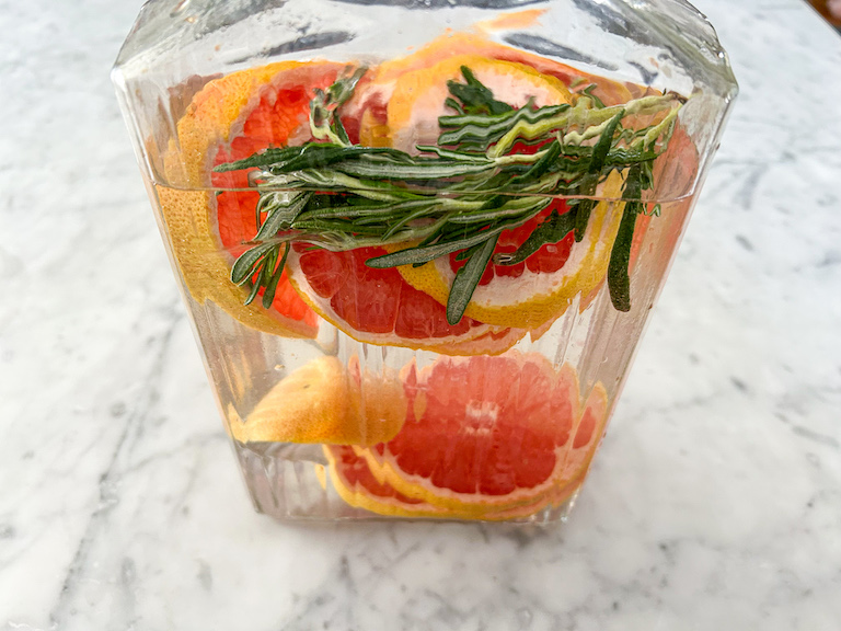 A glass pitcher filled with rosemary, grapefruit, and water