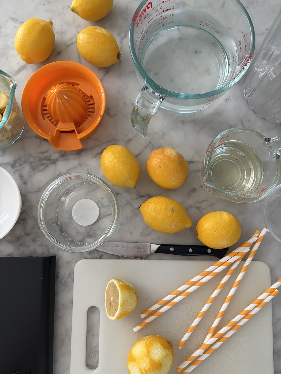 lemons, pitcher of water, scale, cutting board, knife, citrus juicer, and cup of simple syrup on a marble surface