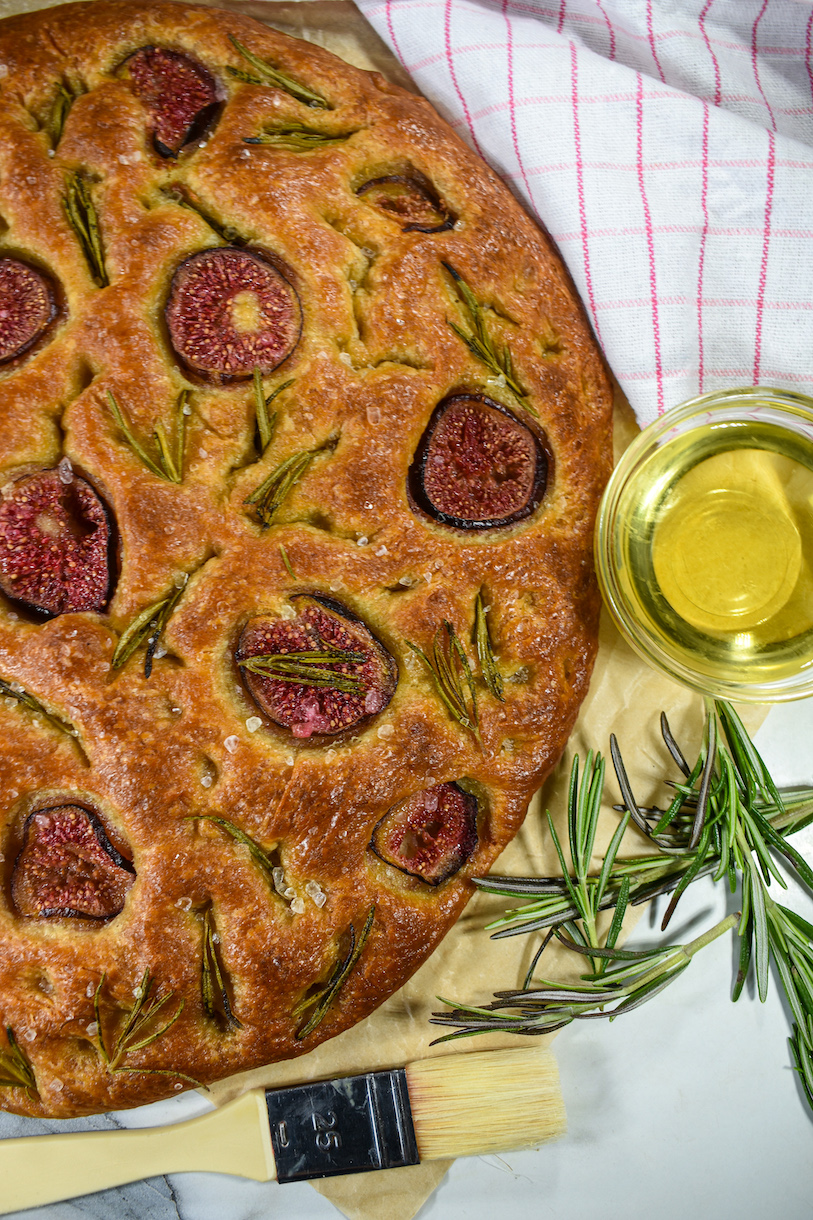 a loaf of fig and rosemary focaccia bread with a dish of olive oil and sprigs of rosemary