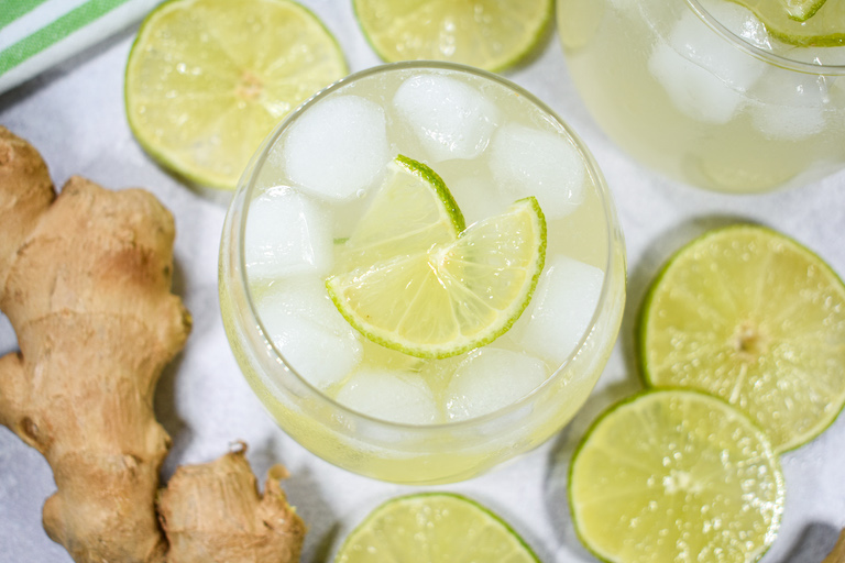 Looking down at a glass of limeade surrounded by slices of lime and a piece of fresh ginger root