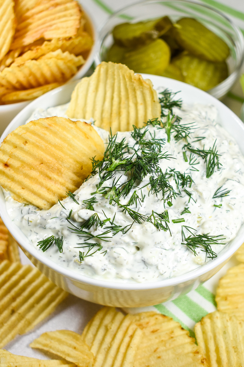 A bowl of creamy dill dip with potato chips and a dish of pickles