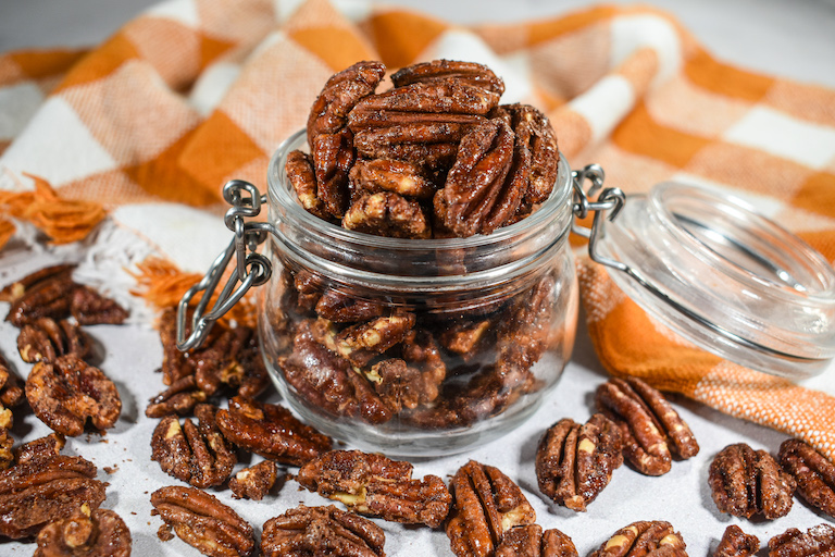 A small glass jar of cinnamon maple pecans with an orange checkered tea towel