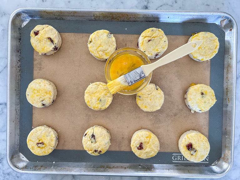 Cranberry scones on a tray before baking, along with a pastry brush and a dish of egg wash