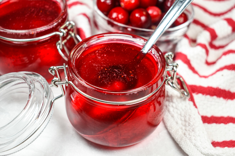 A jar of cranberry syrup with a spoon, red striped towel, and dish of cranberries
