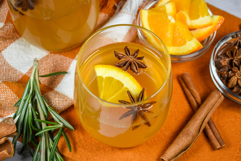 A glass of mulled white wine sitting on an orange tea towel surrounded by cinnamon sticks, orange slices, and sprigs of rosemary