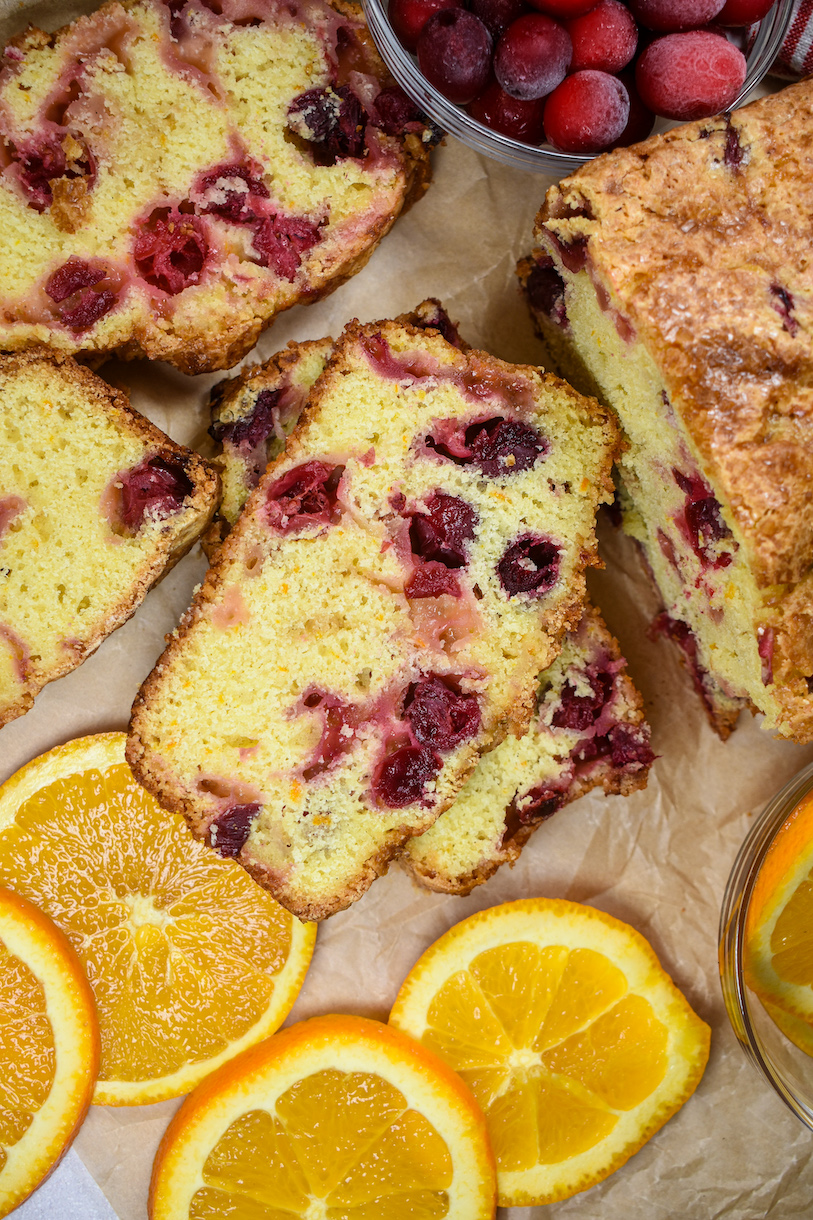 Slices of cranberry orange bread, a dish of cranberries, and slices of fresh orange
