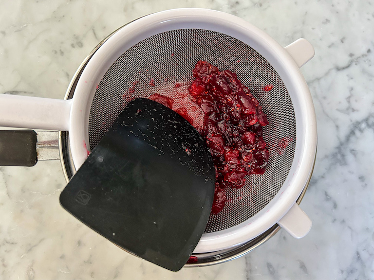 Pushing cranberry syrup through a fine mesh strainer with a bench scraper
