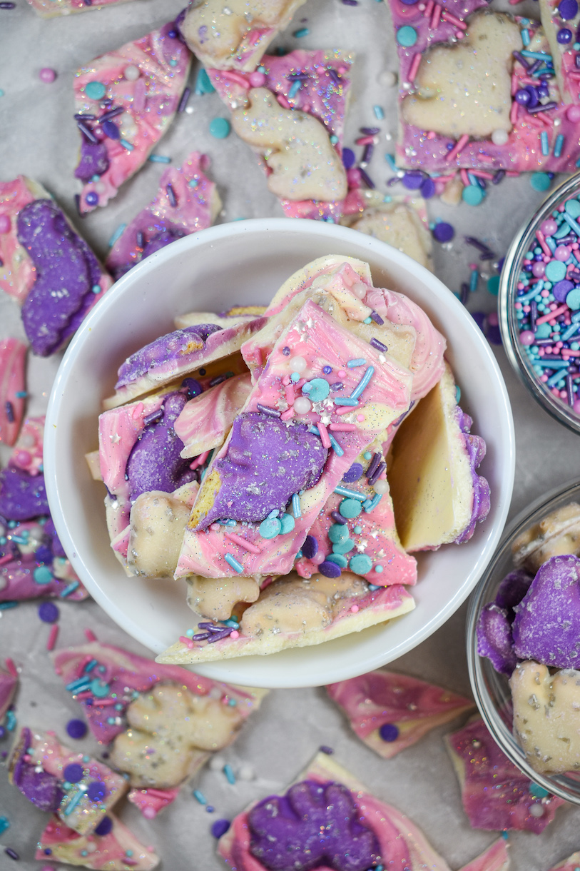 A bowl of white chocolate unicorn bark surrounded by pieces of unicorn candy bark on a white surface