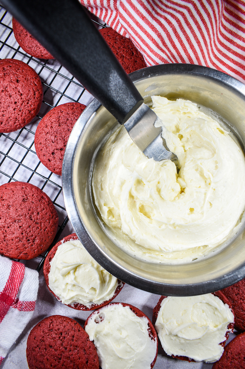 A bowl of cream cheese frosting, small palette knife, and wire rack with red velvet whoopie pies