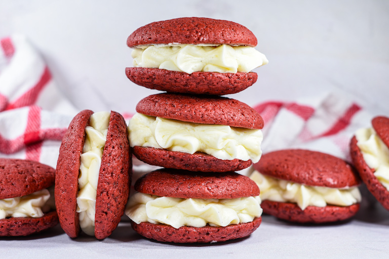 A stack of mini red velvet whoopie pies filled with cream cheese frosting, arranged in front of a white backdrop