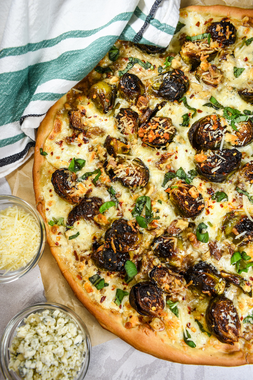 Horizontal shot of a roasted brussels sprout pizza with an green striped towel and bowls of cheese