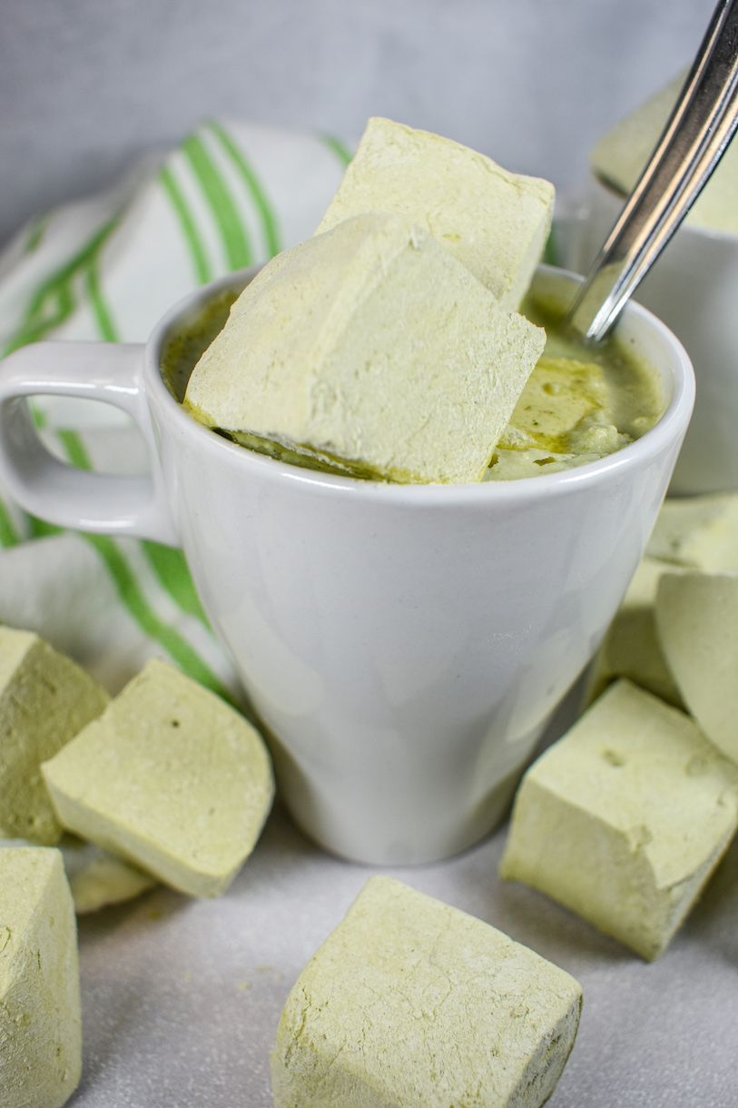 A white mug filled with matcha hot chocolate and matcha marshmallows, surrounded by matcha marshmallows and a green striped towel