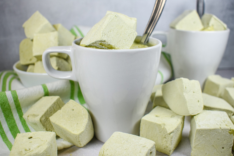 Two mugs of matcha hot chocolate surrounded by matcha marshmallows