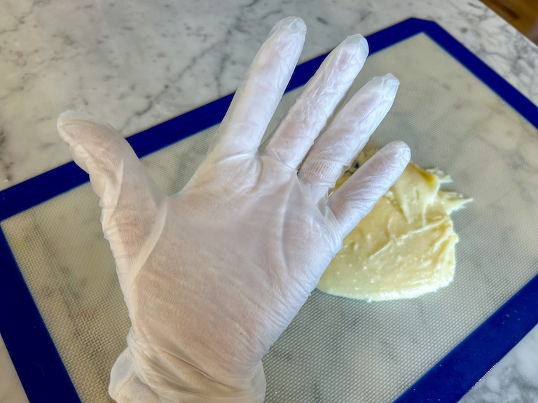 A gloved hand above a silicone mat of candy corn dough, on a marble countertop 
