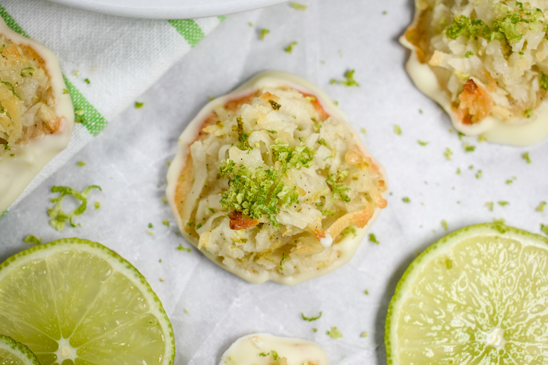 Looking down at a coconut macaroon sprinkled with lime zest, on a sheet of parchment next to slices of fresh lime
