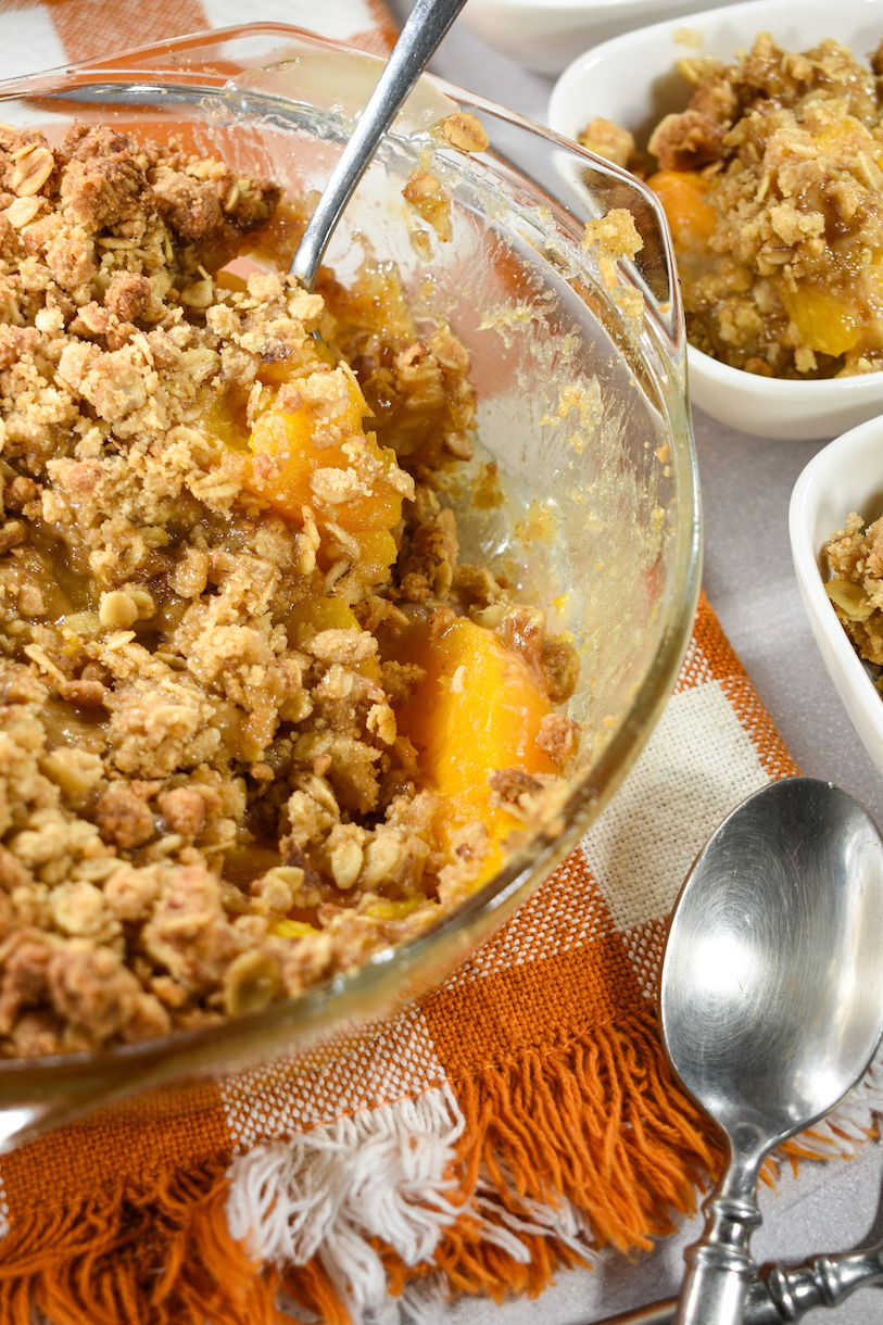 A glass dish of peach crisp on an orange checkered tea towel