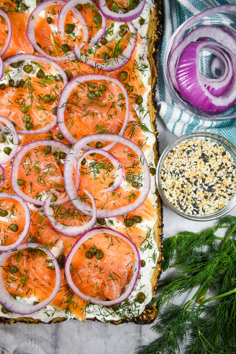 Everything bagel tart on a white surface next to onion rings, dish of everything bagel seasoning, and sprig of dill