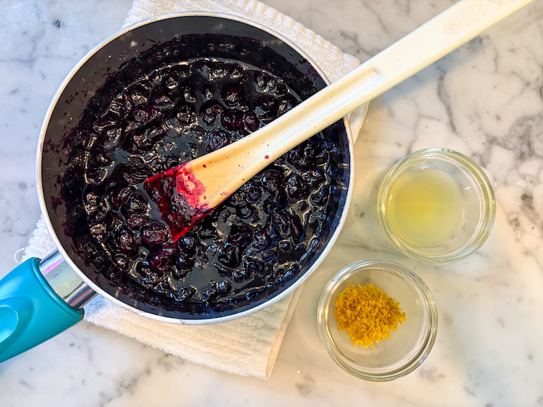 a pan of blueberry compote, dish of lemon juice, and dish of lemon zest on a marble surface