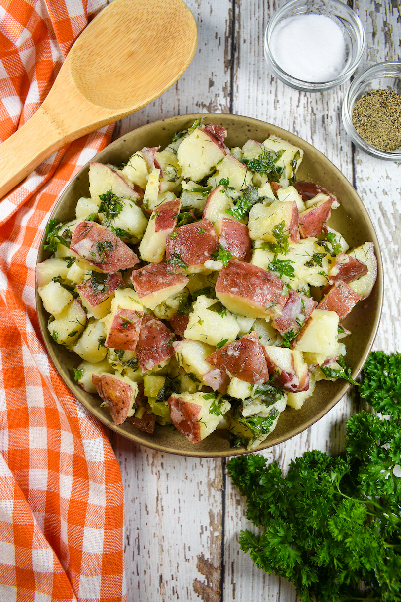 A bowl of red potato salad, checkered tea towel, wooden spoon, and sprigs of fresh parsley