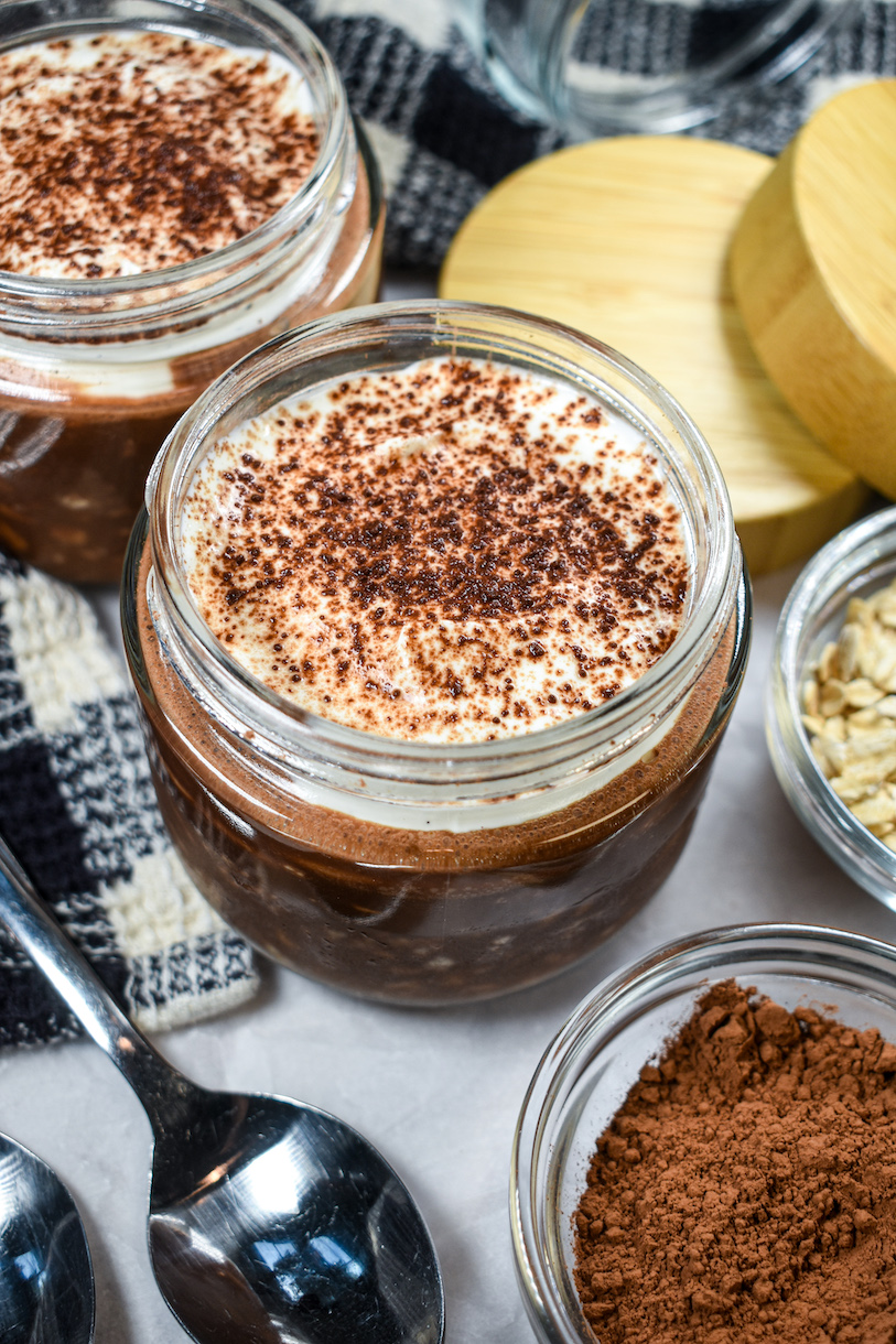 Jars of overnight oats and a checkered towel on a white surface with dishes of cocoa powder and oats