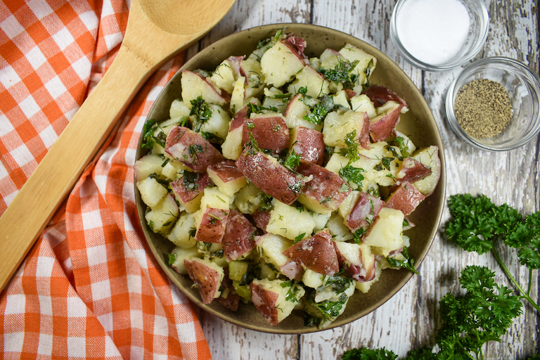 A bowl of red potato salad with a checkered tea towel and wooden spoon
