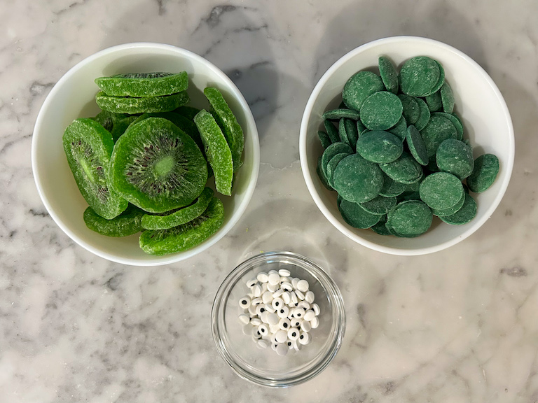 Dish of kiwi, dish of candy melts, and dish of candy eyeballs on a marble surface