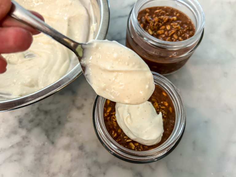 Hand spooning cream topping over jars of overnight oats