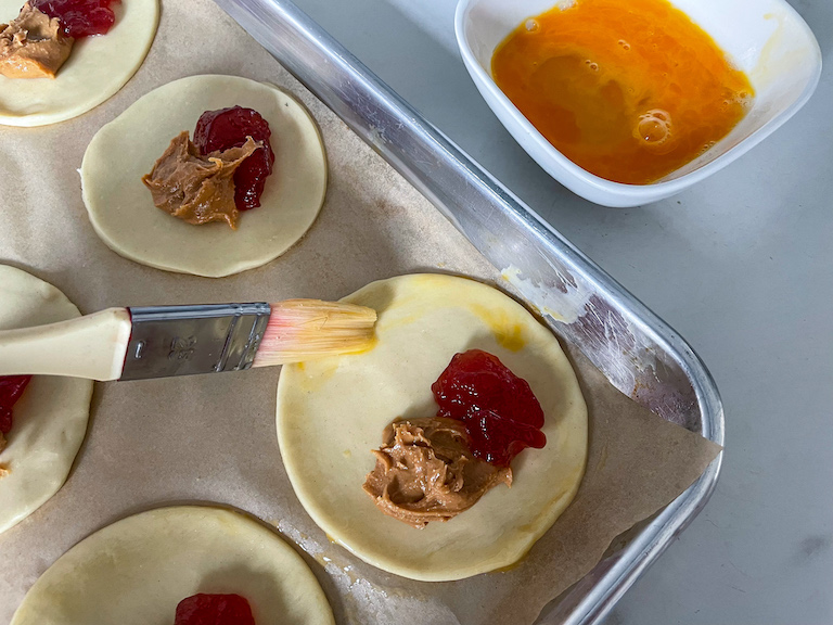 a dish of egg wash, pastry brush, and unbaked peanut butter and jelly hand pies on a tray
