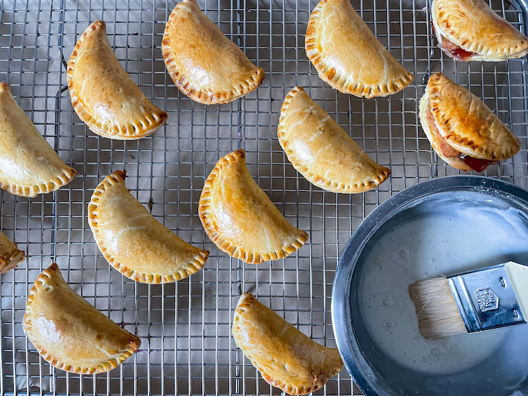 Hand pies on a wire rack with a bowl of glaze and a pastry brush