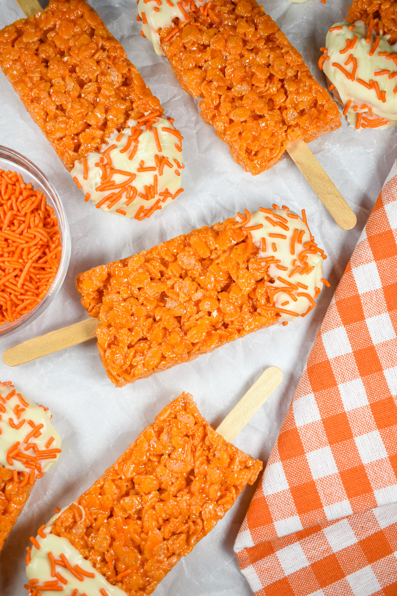 Orange Creamsicle cereal treats arranged on a white surface with a tea towel and dish of sprinkles