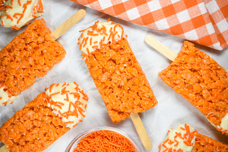 Krispie treat pops arranged on a white surface with a checkered orange tea towel and dish of sprinkles