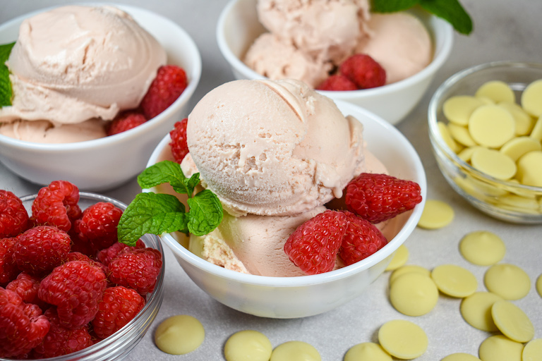 dishes of raspberry ice cream on a white surface with pieces of white chocolate and a dish of fresh raspberries