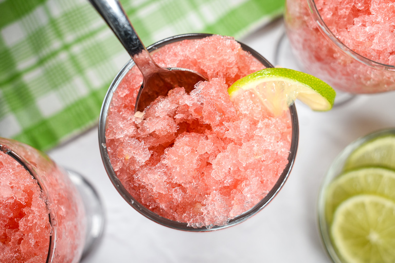 Looking down at a glass of watermelong granita with a lime slice and a metal spoon