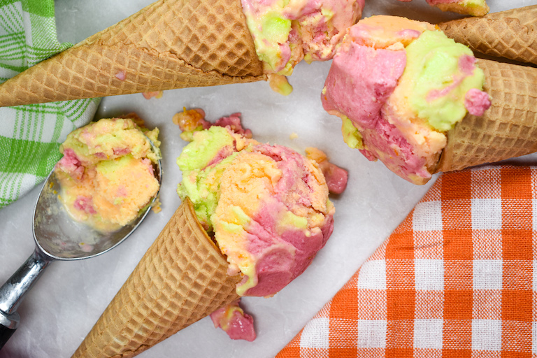 Ice cream cones with scoops of rainbow sherbet, arranged on a white surface with colorful tea towels