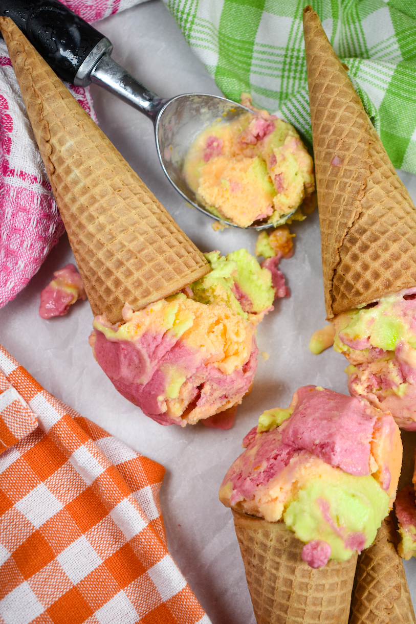 Cones of homemade rainbow sherbet on a white surface with colorful checkered tea towels