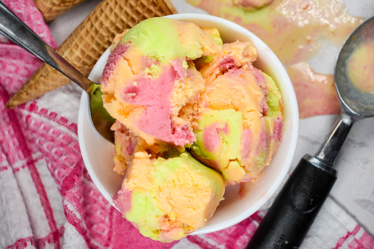 A bowl of homemade rainbow sherbet with a pink towel, ice cream cones, and an ice cream scoop